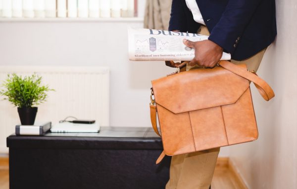Finance apps: Man leaning against a wall holding a tan leather briefcase and a newspaper